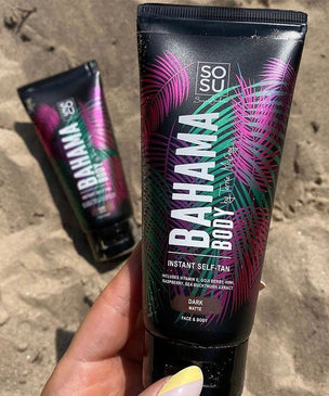 A hand holding a Bahama Body instant self-tan tube in a dark shade, with vibrant palm leaf design, against a sandy beach background. Another tube is visible in the background.