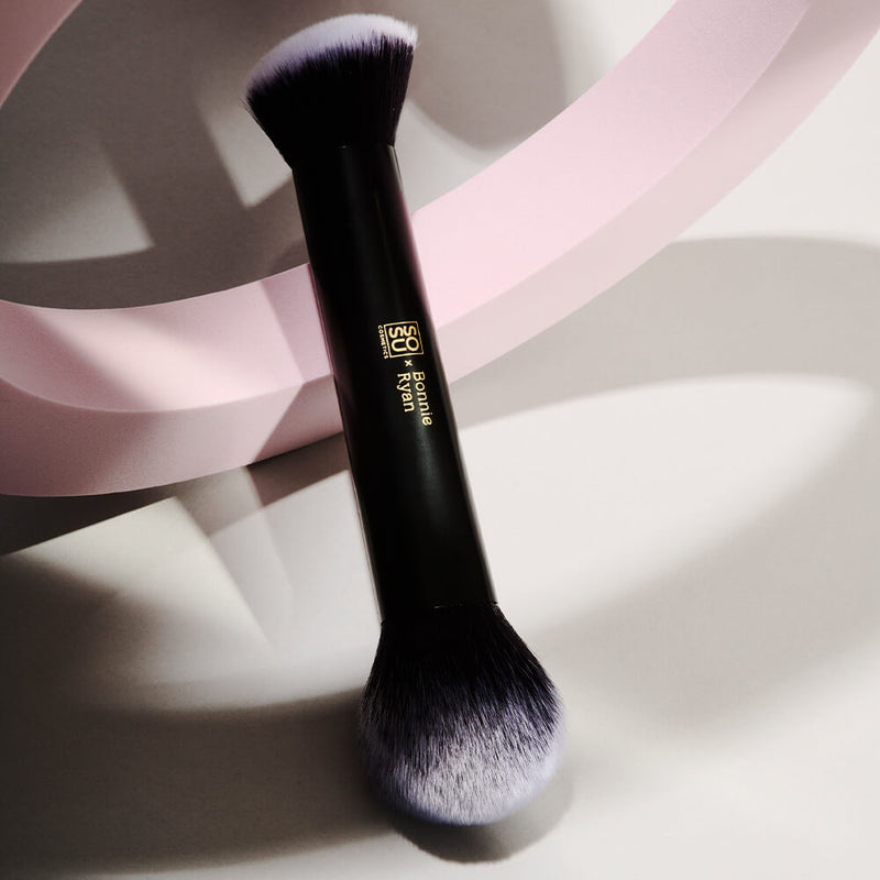 Close-up view of the SOSU x Bonnie Ryan makeup brush, showcasing its duo-toned bristles. The brush lies elegantly against a backdrop of soft pink curves, casting gentle shadows and emphasizing its sleek black handle with the signature gold branding.