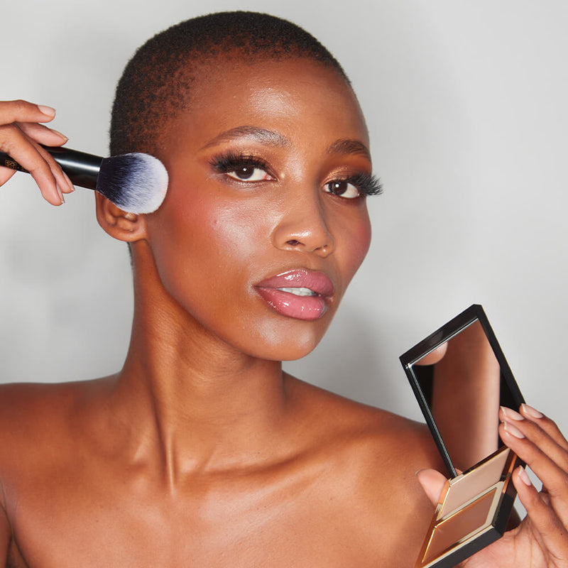A model with a radiant complexion and short hair confidently holds the SOSU x Bonnie Ryan dual-bristled makeup brush to her cheek, demonstrating its use. Her luminous makeup is accentuated by the brush, and she also presents a sleek, rectangular makeup compact in her other hand against a light background.
