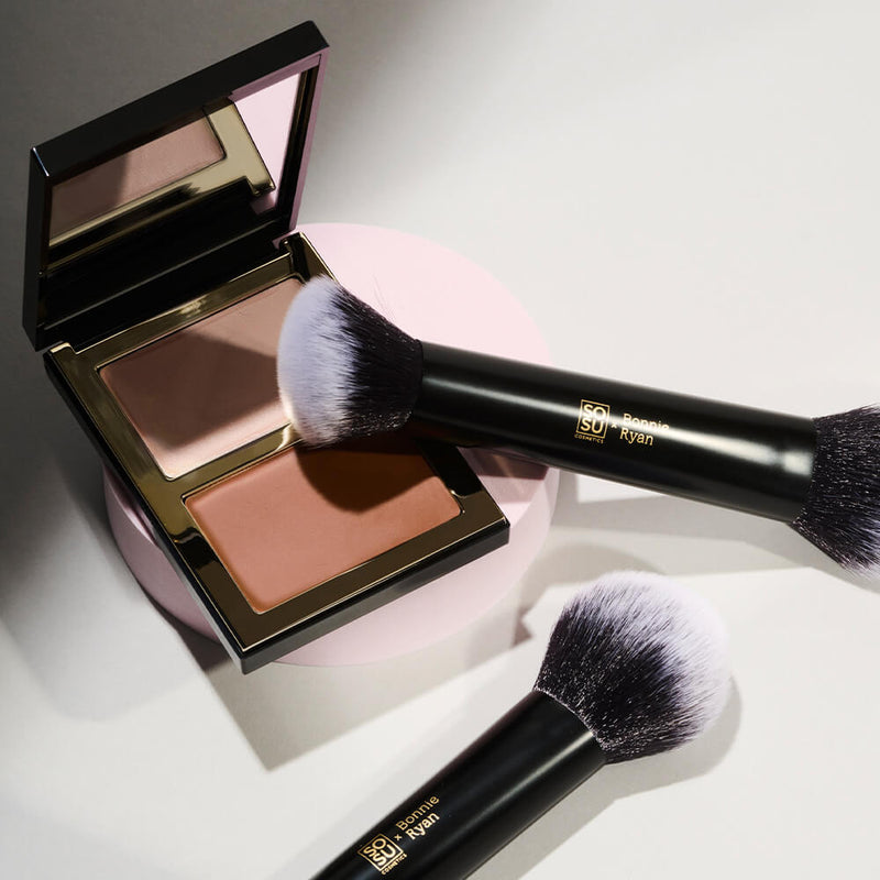 A close-up view of the SOSU x Bonnie Ryan dual-bristled makeup brush with its brand details embossed on the handle, resting against an open makeup compact. The compact showcases a two-toned blush palette with a reflective casing, all positioned on a pastel pink circular backdrop with contrasting shadows.