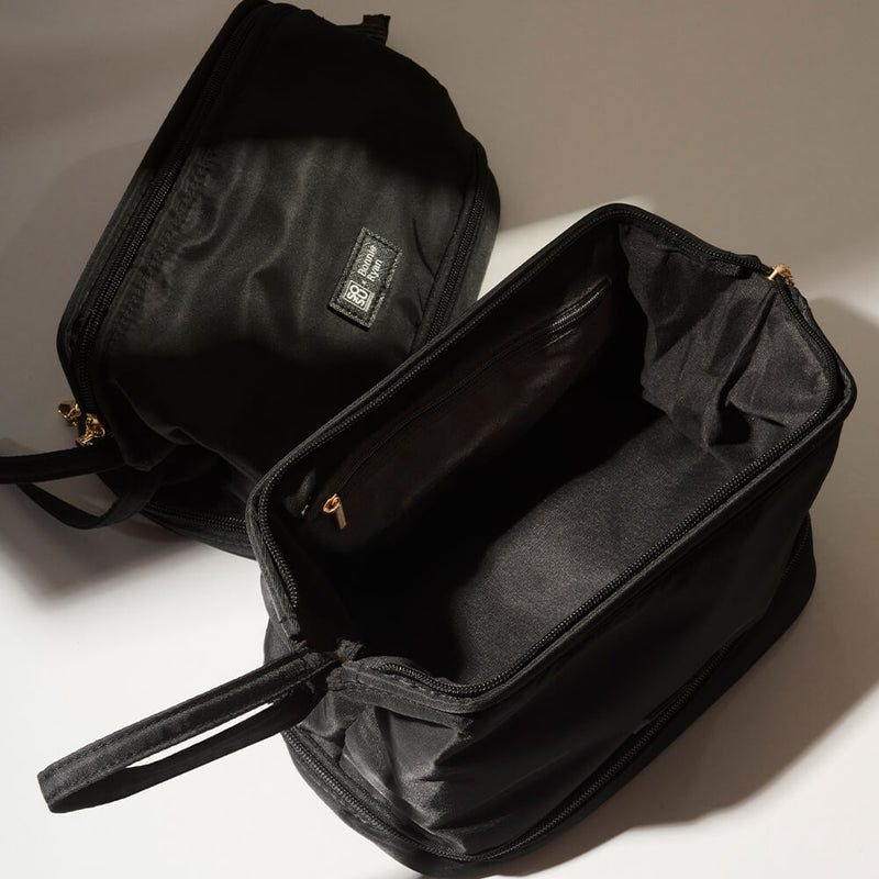 Two SOSU x Bonnie Ryan black makeup bags set against a soft-lit backdrop. One bag stands vertically, showing its subtly embossed brand label and a side zippered compartment. The other bag lies horizontally with its main compartment partly unzipped, revealing its spacious black interior and an elegantly attached handle. Both bags showcase gold-tone zipper pulls, emphasizing their premium design.