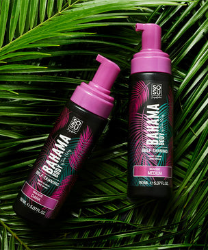 Two bottles of 'Bahama Body Mousse' in vibrant pink packaging, nestled among lush green palm leaves.