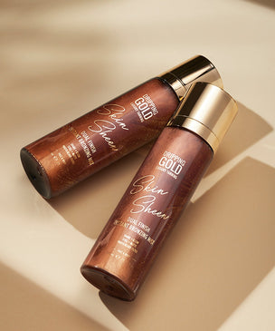 Two 'Dripping Gold Skin Sheen' products with a radiant copper hue, elegantly placed against a soft-lit neutral background, representing the Body Finish & Glow collection.