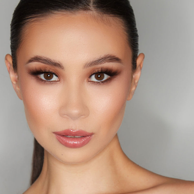 Close-up portrait of a model showcasing a flawless makeup look. Her eyes are accentuated with deep, smoky eyeshadow and lush eyelashes, while her skin glows with a radiant finish. Subtle pink lip gloss adds the finishing touch to her polished appearance.