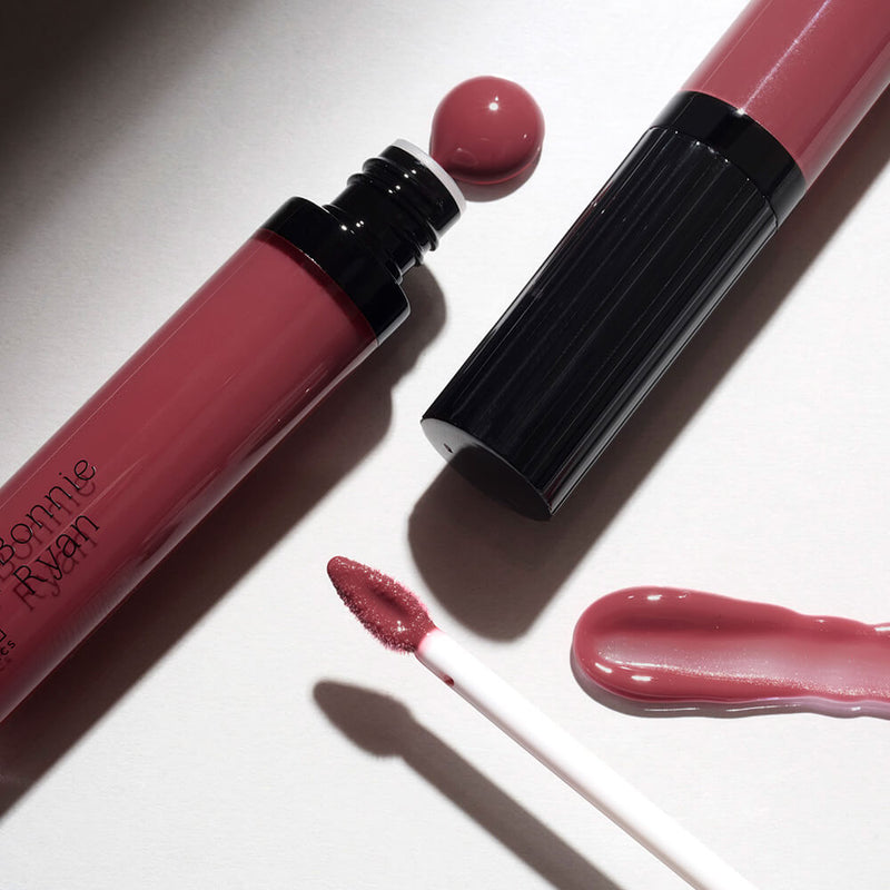 Close-up of a SOSU x Bonnie Ryan pink lip gloss tube with its applicator dipped in the vibrant product, next to a smeared gloss sample. The scene is illuminated by a soft light, creating gentle shadows on the surface.