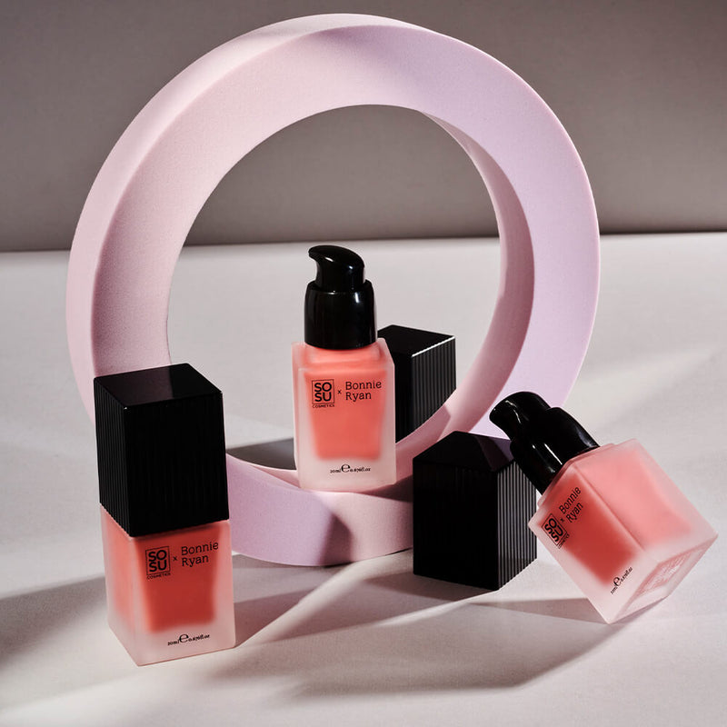 Elegant display of the SOSU x Bonnie Ryan Liquid Blush bottles, nestled inside a curved lavender-hued structure. Each blush bottle, featuring a black pump and label, exudes a soft coral tint, set against a neutral background, casting gentle shadows.