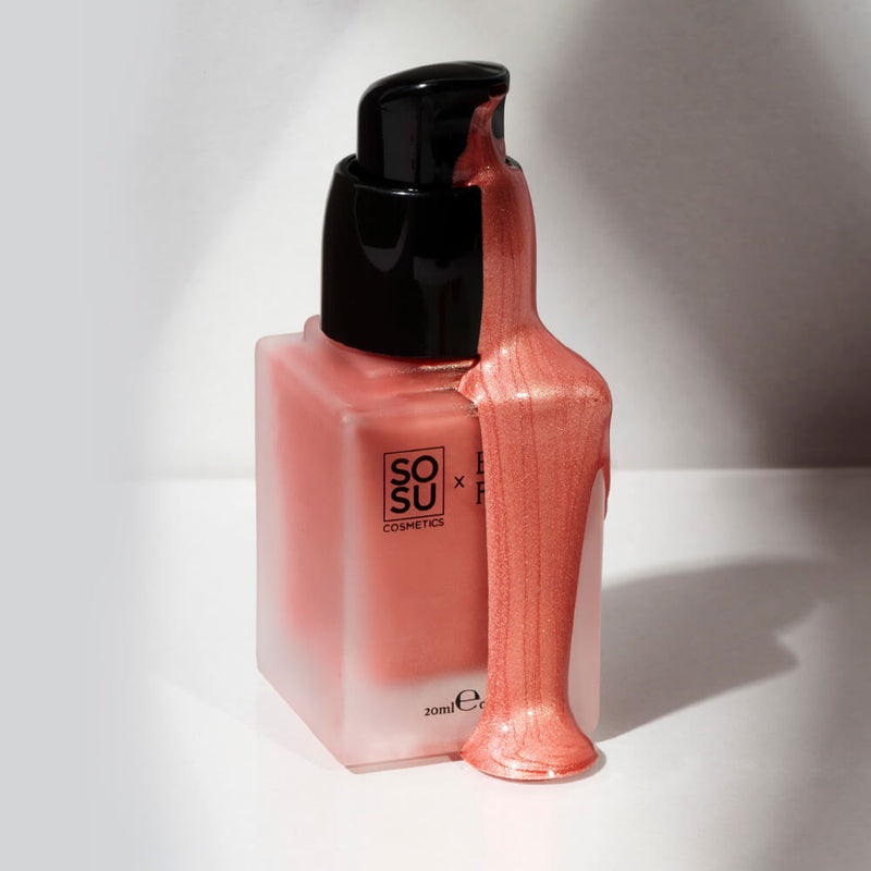 Close-up of a SOSU x Cosmetics liquid blush bottle with a luxurious spill of shimmering coral-hued product cascading down its side. The bottle's label and a 20ml indicator are clearly visible, all illuminated under soft lighting against a muted background.